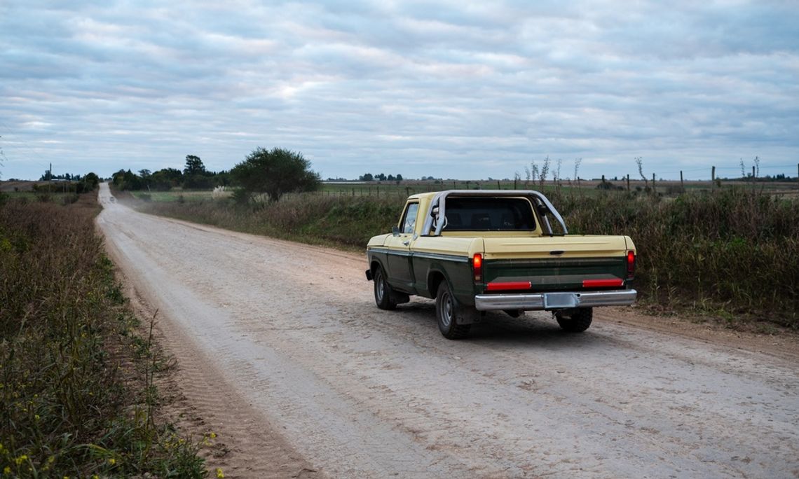 Why Rural Roads Can Be Tough on Vehicle Frames A green and cream vintage pickup truck driving along a wide dirt road bordered by grassy fields beneath a cloudy sky.
