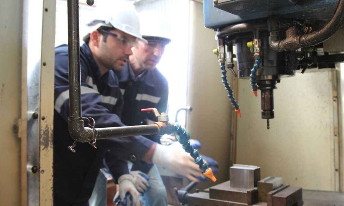 Two Caucasian men wearing hard hats and safety goggles observe a large machine surrounded by metal walls.