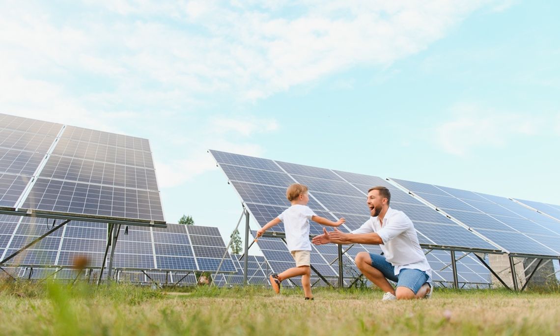 A man is kneeling down as a small child runs into his open arms. They are outside, in front of a set of solar panels.