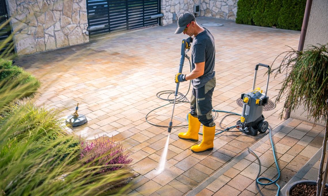 Why Some Outdoor Cleaning Projects Take Longer Than Expected A man wearing yellow rubber boots pressure washes a brick patio beside landscaping, with cleaning equipment nearby.