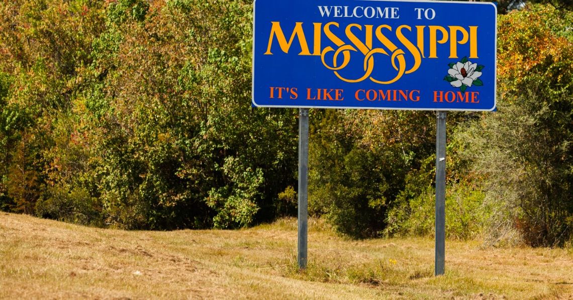A big blue road sign state sign along the highway. The sign reads "Welcome to Mississippi, it's like coming home."