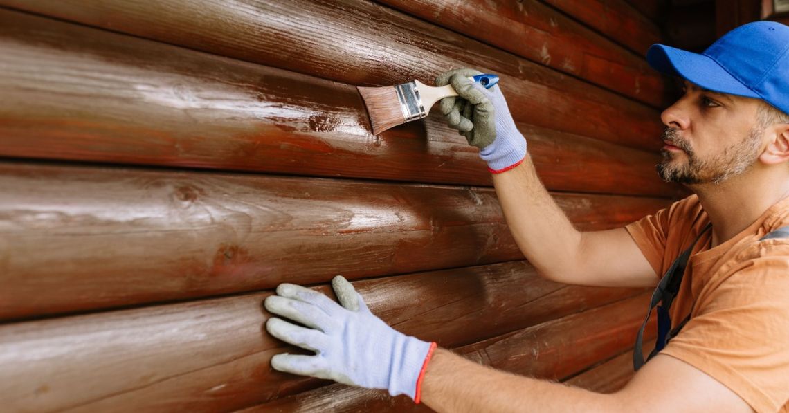 Why You Shouldn’t Paint the Exterior of a Log Cabin A man wearing gloves and a blue cap brushing paint onto a glossy brown log cabin wall while steadying a lower log.