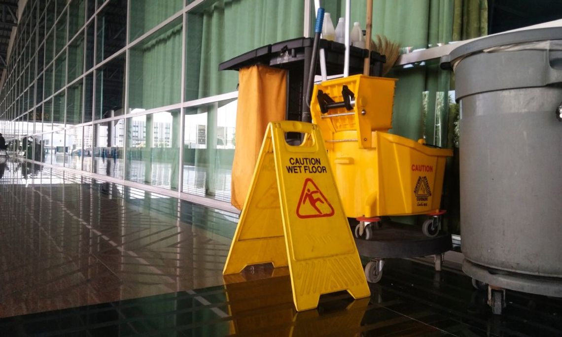 A business hallway with a cleaning supply cart and a wet floor sign. There is a yellow mop system and a trash can.